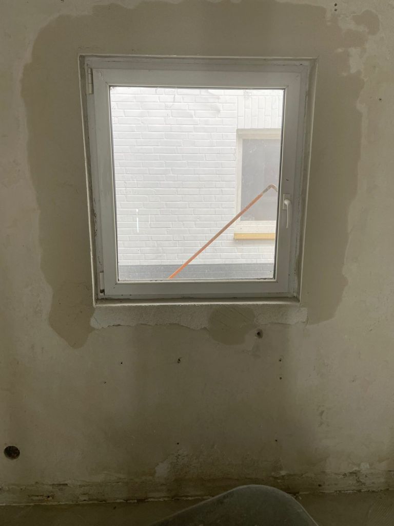 Wandfläche um ein Fenster mit dunkel abgezeichneten Feuchterändern rings um die Laibung, Putz teilweise abgetragen